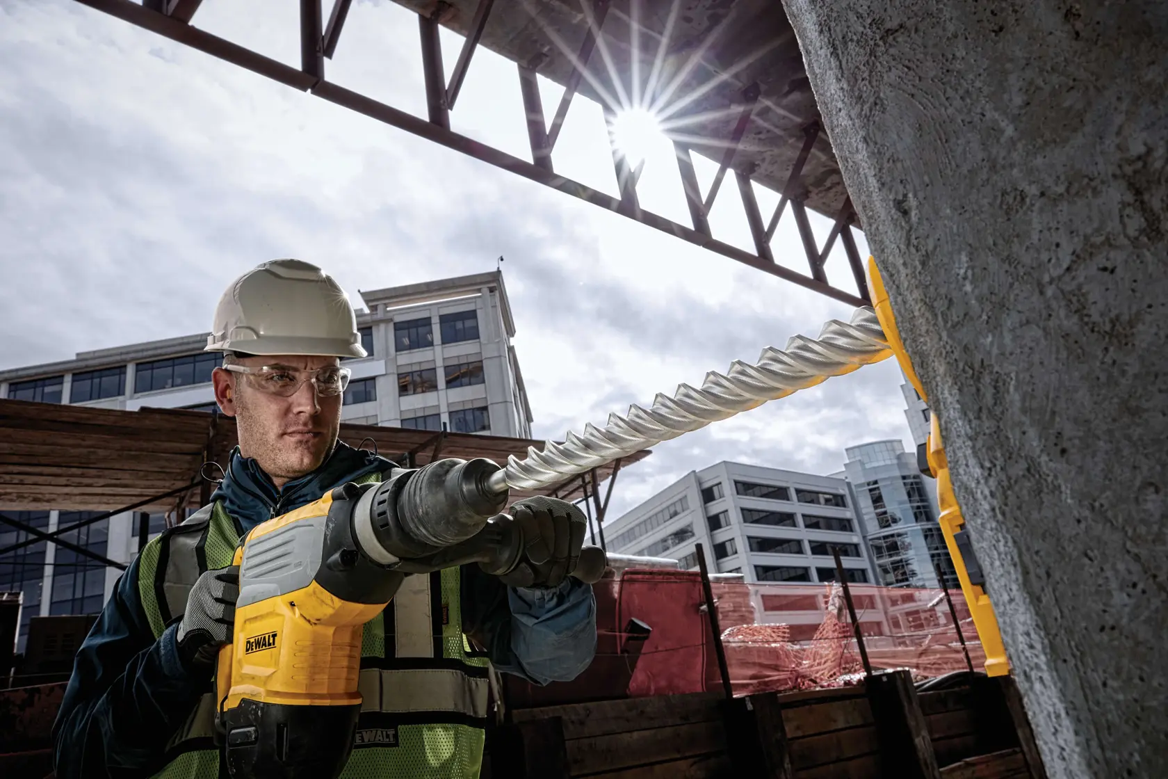 Worker uses DEWALT drill with DT60811 bit on concrete at urban construction site; modern buildings in background.