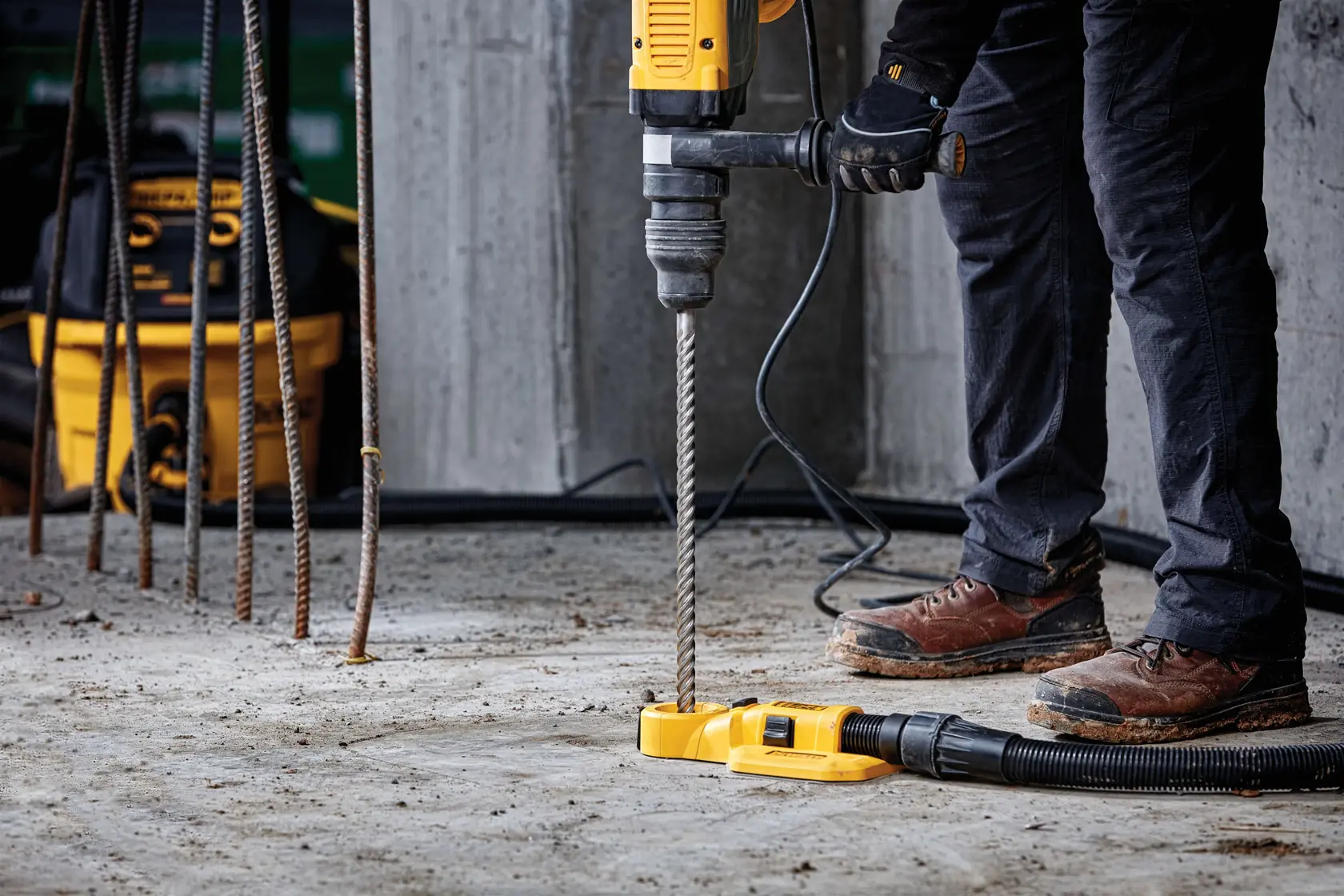 Person uses yellow DEWALT drill with dust extractor to drill into concrete at construction site.