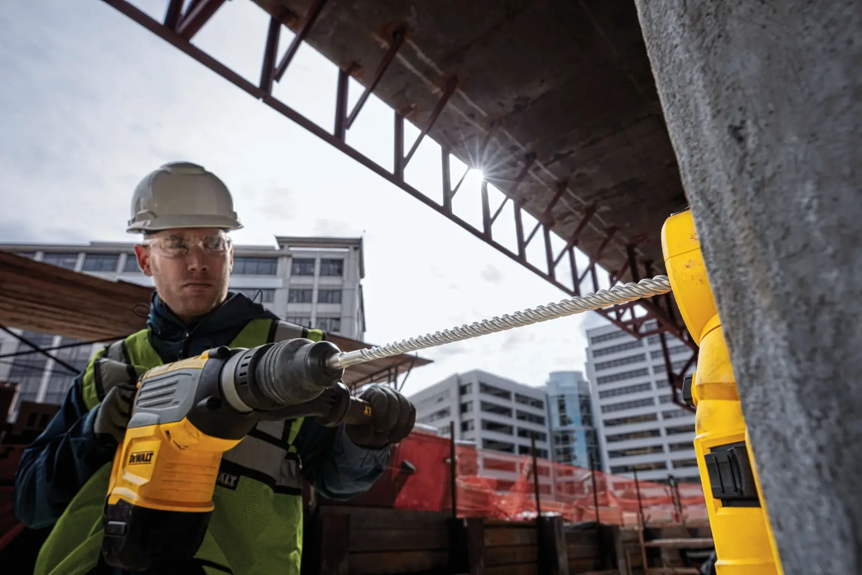 Worker uses a DEWALT drill at a construction site with buildings and safety gear visible in the background.