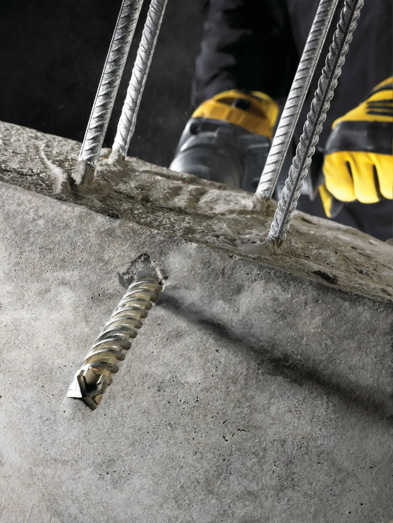 Close-up of a DEWALT drill bit boring into concrete with metal rebar and gloved hands visible.