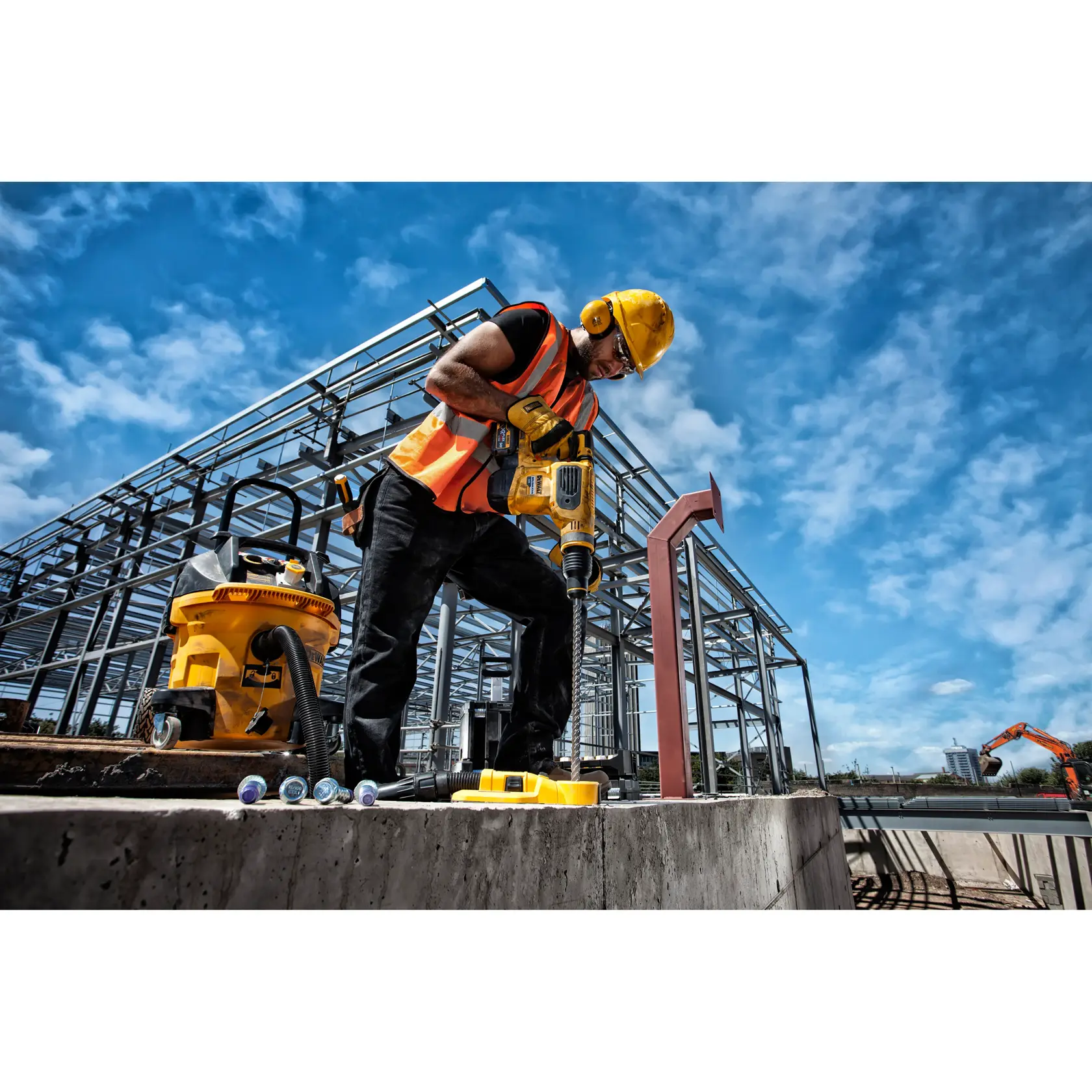 Worker uses DEWALT drill at construction site with steel framework and equipment under a blue sky.