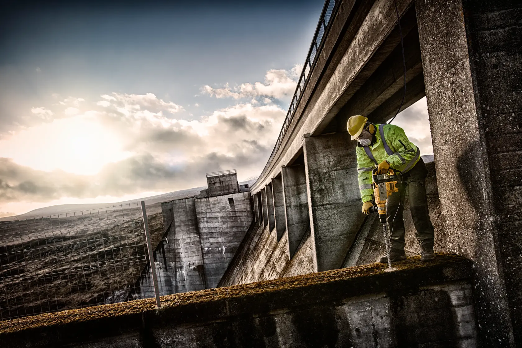 Worker uses DEWALT tool to drill concrete on a large dam structure under a cloudy sky.