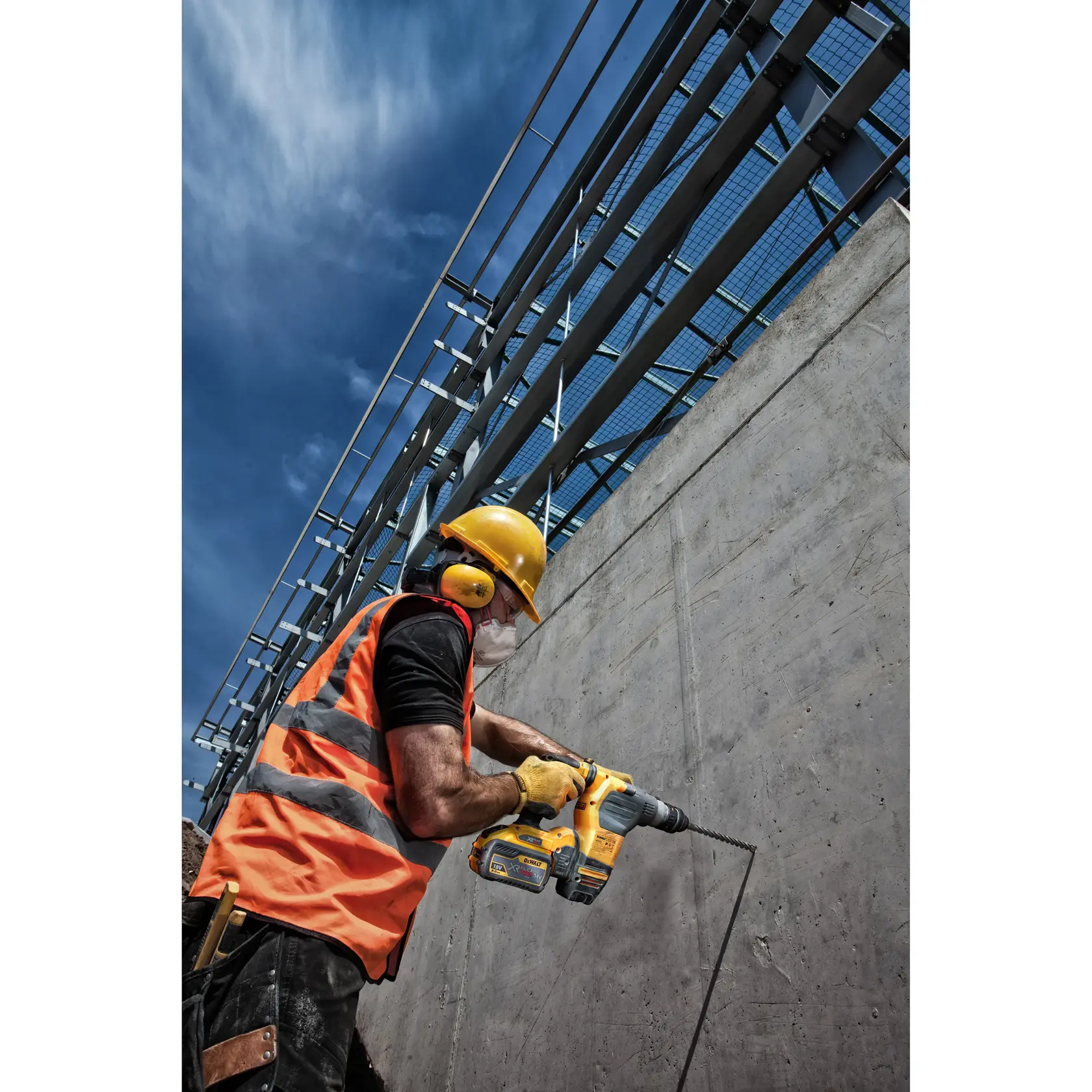 Worker in safety gear drills concrete wall with yellow DEWALT tool at construction site under blue sky.