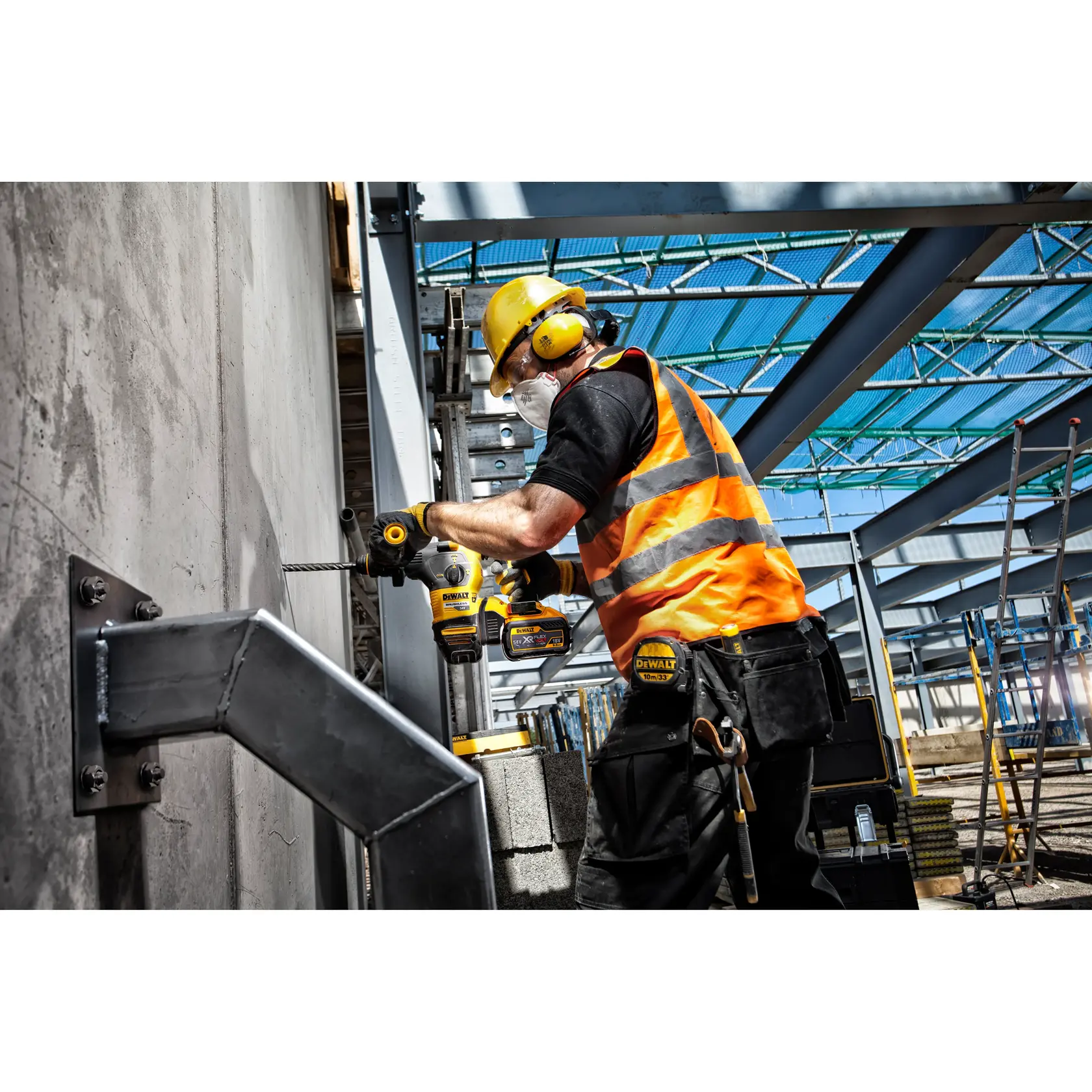 A construction worker wearing a yellow hard hat, ear protection, safety glasses, and an orange reflective vest is using a DEWALT power drill on a concrete wall at a construction site with steel beams overhead.