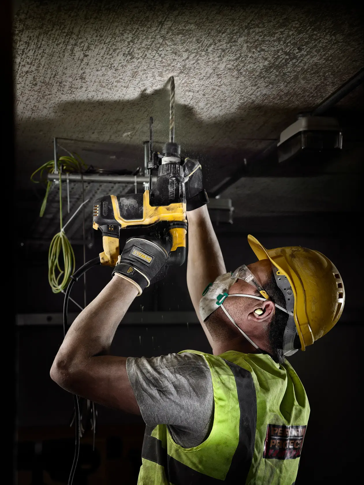 Worker in safety gear uses Dewalt drill to make a hole in the ceiling at a construction site.