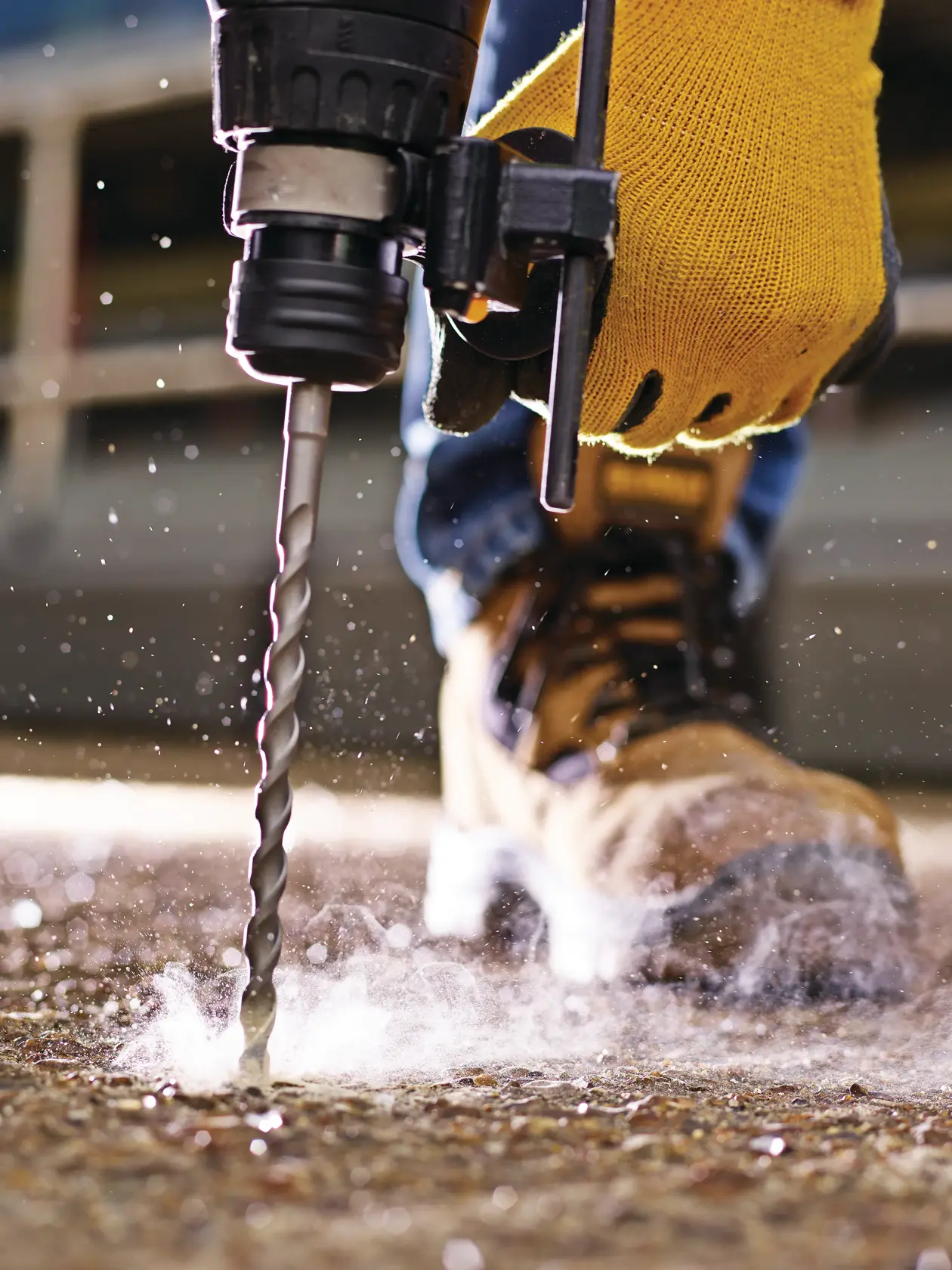Close-up of a gloved hand using a drill bit on concrete, with dust and work boot visible.