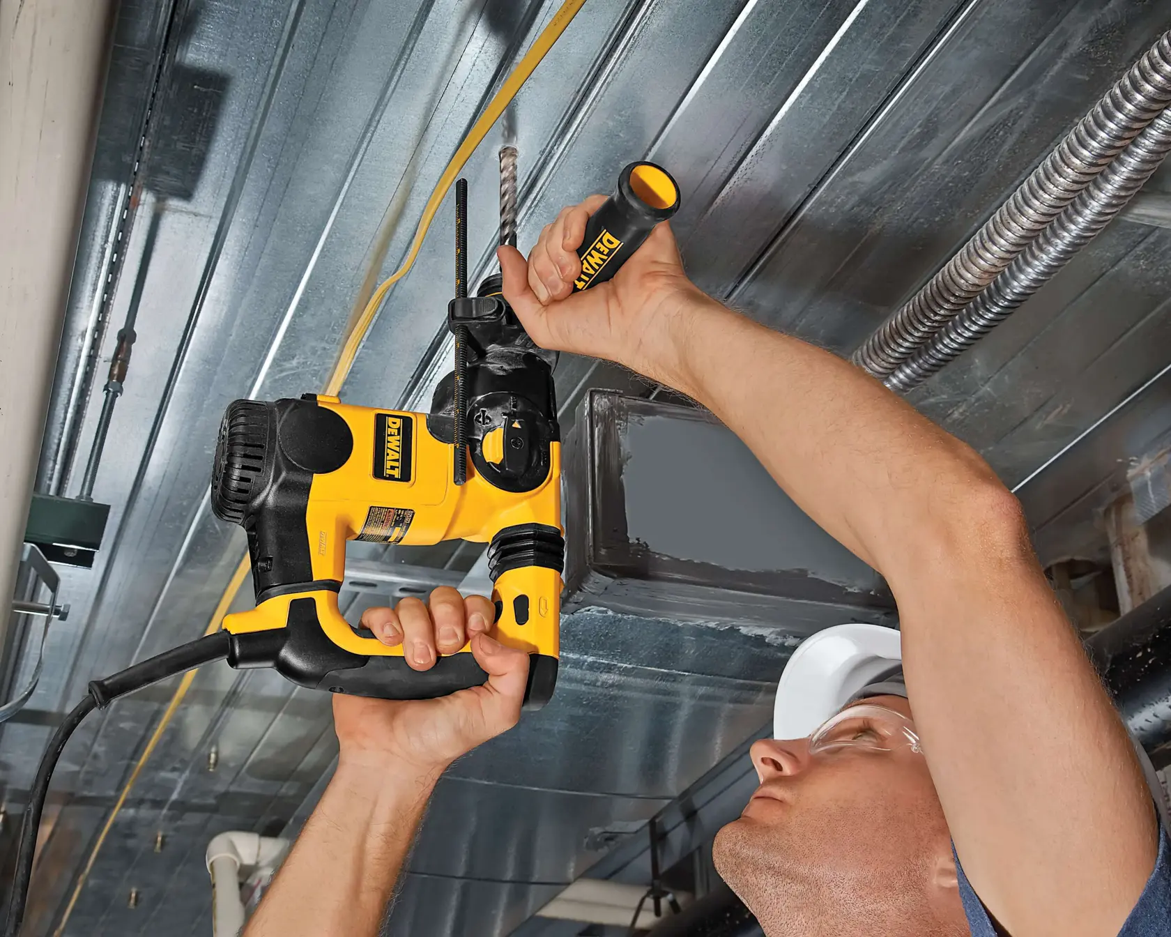 A person using a DEWALT rotary hammer drill to drill into a metal ceiling. The drill is yellow and black, and the person is holding the tool with both hands while working overhead.