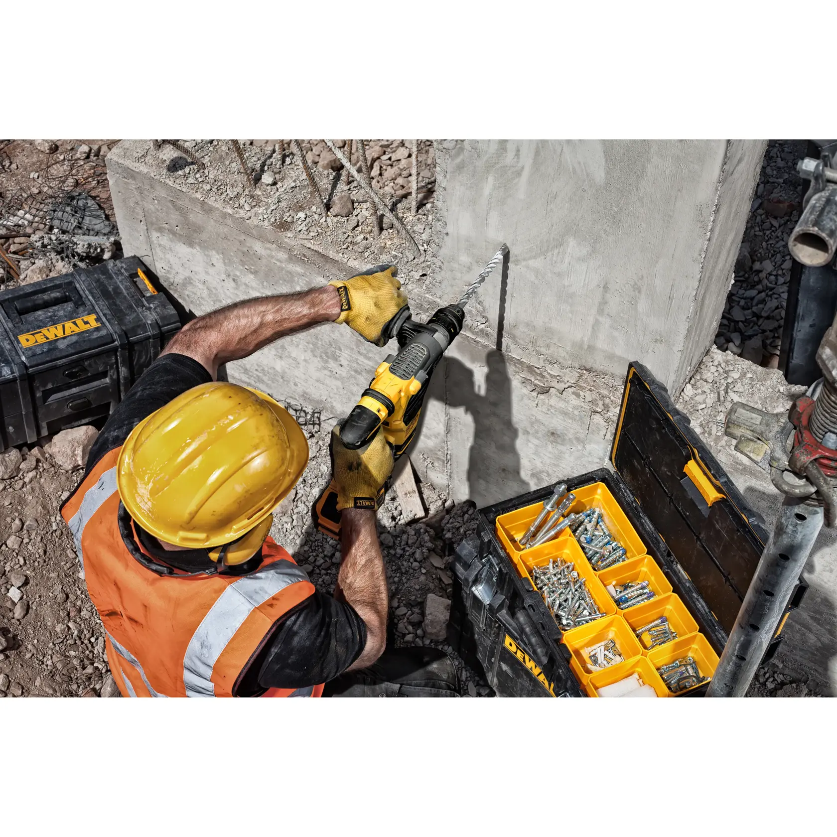 Worker drills into concrete with DEWALT tool, using a screw kit and wearing safety gear at a construction site.