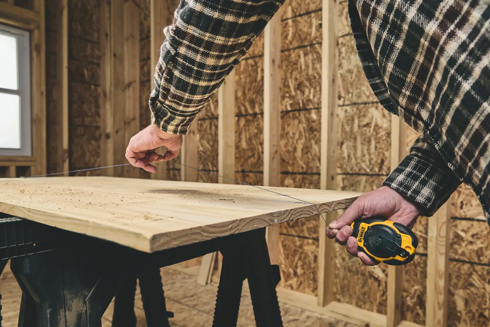 A person using a DEWALT chalk line tool to mark a straight line on a wooden board in a workshop or construction setting.