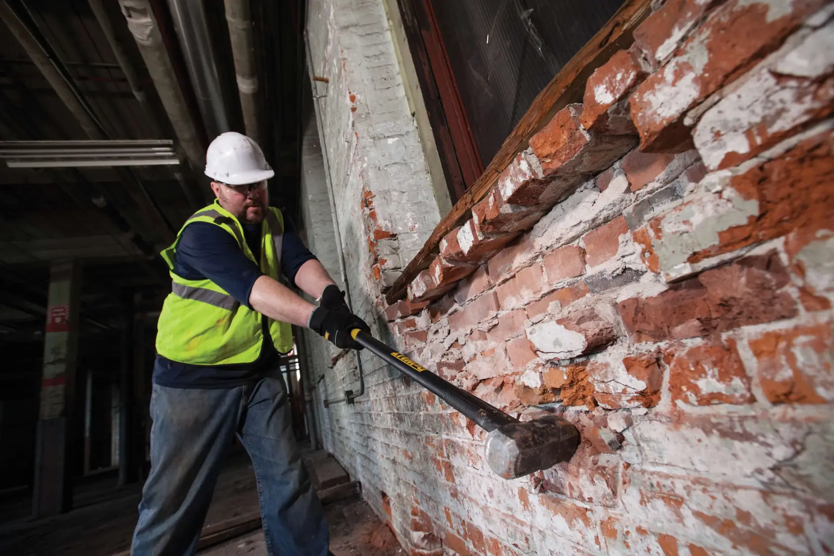 A person wearing a safety vest and hard hat is using a DEWALT sledgehammer to demolish a brick wall inside an industrial building.